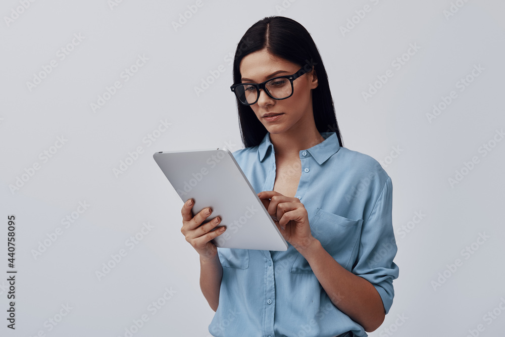 Attractive young woman in eyewear using digital tablet while standing against grey background Attractive young woman in eyewear using digital tablet while standing against grey background