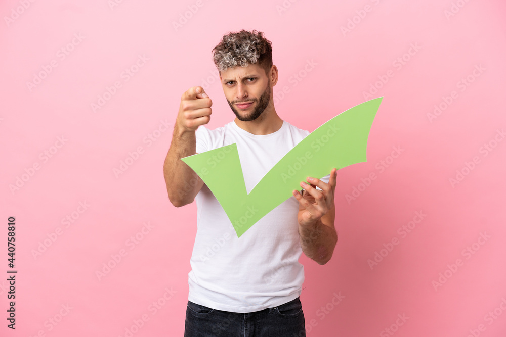 Young handsome caucasian man isolated on pink background holding a check icon and pointing to the front Young handsome caucasian man isolated on pink background holding a check icon and pointing to the front