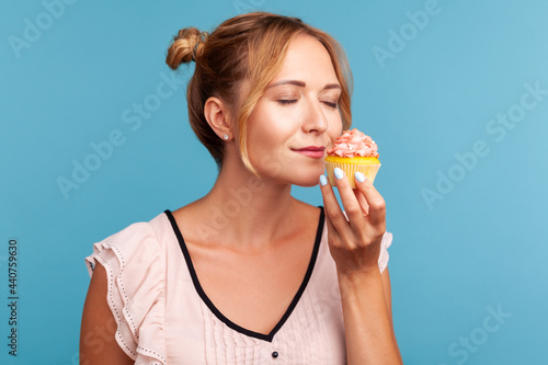 Portrait of beautiful blonde female with hair buns holding cake in hand sand enjoying tasty smelling with closed eyes, having desire to eat dessert. Indoor studio shot isolated on blue background.