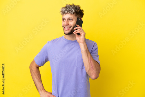 Young caucasian man using mobile phone isolated on yellow background posing with arms at hip and smiling