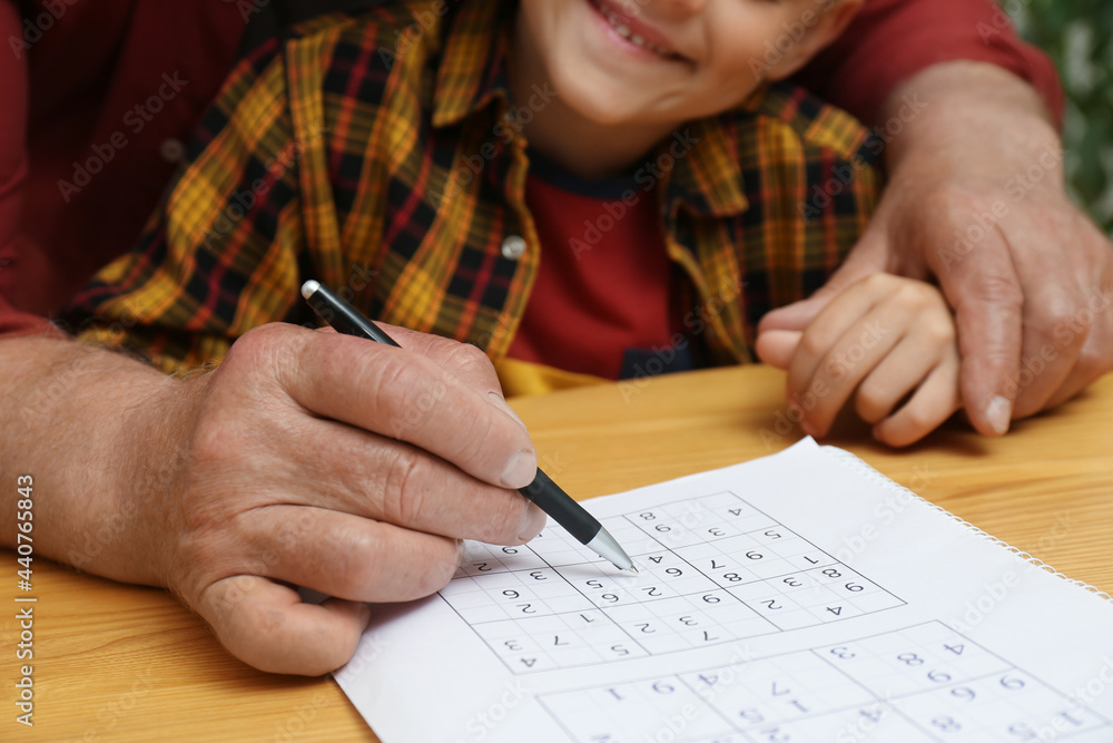 Little boy with his grandfather solving sudoku puzzle at wooden table, closeup Little boy with his grandfather solving sudoku puzzle at wooden table, closeup