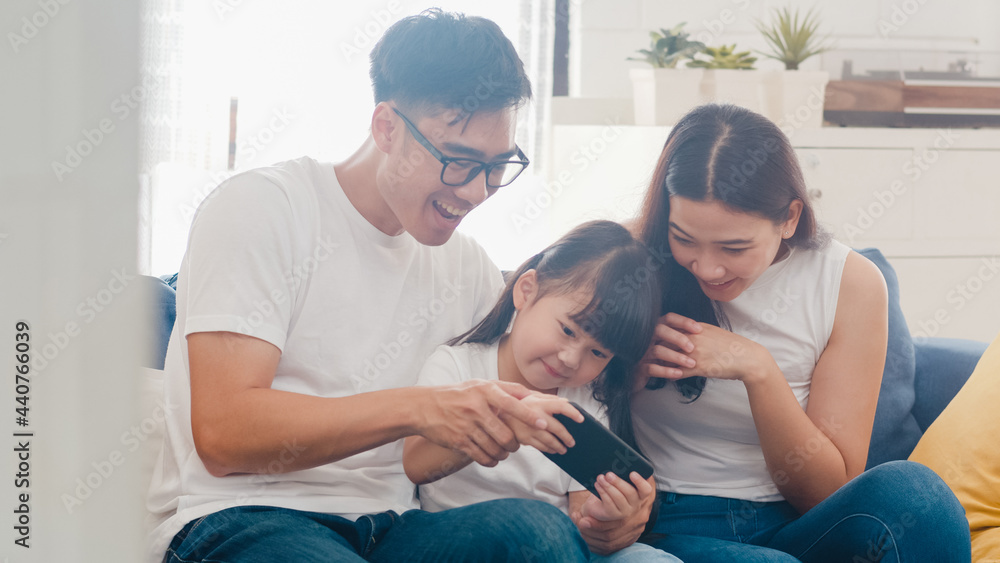 Fototapeta premium Happy Asian family dad, mom and daughter playing funny game online on smartphone sitting sofa in room at house. Self-isolation, stay at home, social distancing, quarantine for coronavirus prevention.