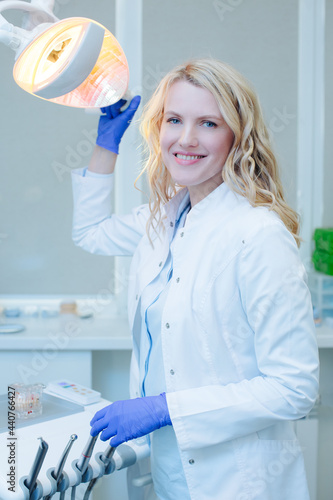 Smiling dentist in a white coat and blue gloves, holding a dental instrument stay at the dental chair in the light of a special lamp