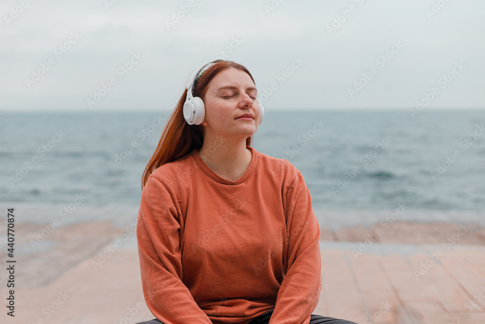 Beautiful caucasian young woman meditates with wireless headphones on the beach near the sea