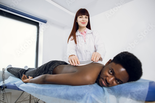 Osteopathy, alternative medicine, back pain relief concept. Physiotherapy and sport injury rehabilitation. Young Caucasian woman masseur making back massage of African man with steel scraper