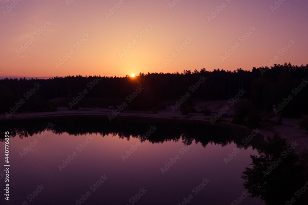 The sun shines from behind the silhouettes of the pine forest, which is reflected in the mirror water of the lake