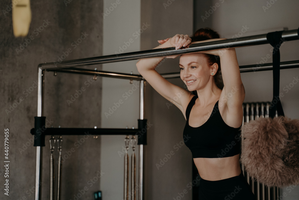 Young happy sportive ginger woman leaning on bar of pilates machine Young happy sportive ginger woman leaning on bar of pilates machine