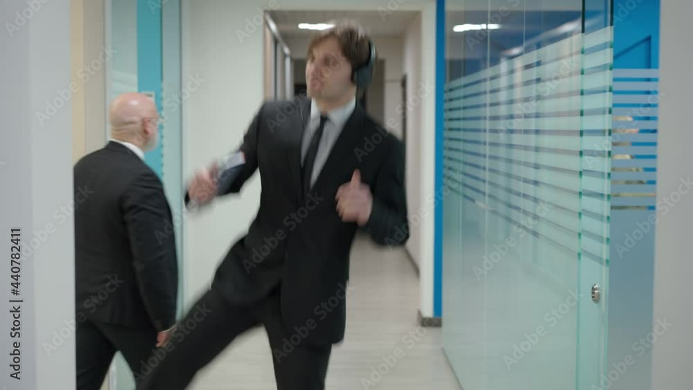 Portrait of joyful happy young handsome Caucasian man in headphones walking in office hallway listening to music dancing as colleagues passing in corridor. Business success and joy concept Portrait of joyful happy young handsome Caucasian man in headphones walking in office hallway listening to music dancing as colleagues passing in corridor. Business success and joy concept