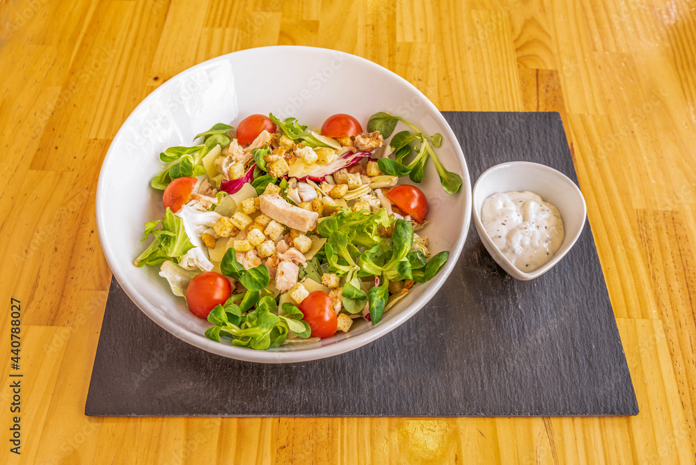 Popular cesar salad with sauce on the side in a white bowl, lamb's lettuce sprouts, roasted chicken pieces, fried croutons, lettuce and cherry tomatoes, all in a white bowl Popular cesar salad with sauce on the side in a white bowl, lamb's lettuce sprouts, roasted chicken pieces, fried croutons, lettuce and cherry tomatoes, all in a white bowl