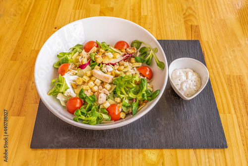 Popular cesar salad with sauce on the side in a white bowl, lamb's lettuce sprouts, roasted chicken pieces, fried croutons, lettuce and cherry tomatoes, all in a white bowl
