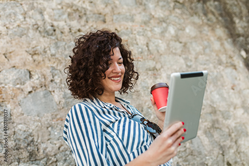 Young woman with curly hair drinking coffee and using her digital tablet while standing outside © Mediteraneo