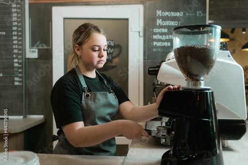 A barista grinds beans in a coffee grinder in a cafe and holds a portafilter. Sad girl waiter with a portafilter in his hands. The process of preparing coffee for a coffee machine.