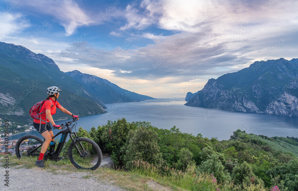 nice senior woman with elctric mountain bike resting on Monte Brione and enjoying the awesome view over Garda Lake between Riva del Garda and Torbole - obrazy, fototapety, plakaty 