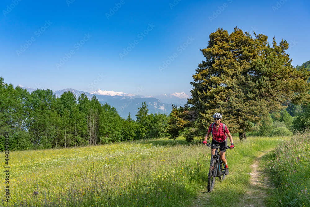 nice, active senior woman riding her electric mountain bike at the slopes of Monte Velo in the Sarca Valley with Monte Adamello in background, Garda lake mountains, Trentino, Italy 
