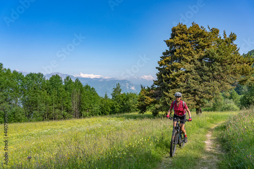 nice, active senior woman riding her electric mountain bike at the slopes of Monte Velo in the Sarca Valley with Monte Adamello in background, Garda lake mountains, Trentino, Italy 