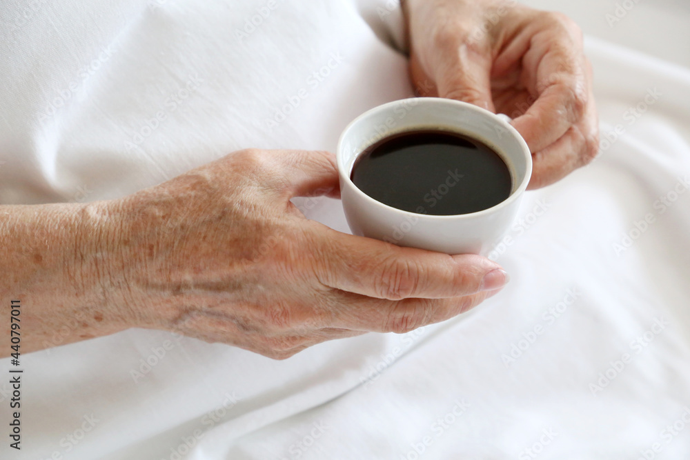 Elderly woman with white cup of black coffee in wrinkled hands sitting in a bed. Concept of fresh morning, energy and enjoying life at retirement Elderly woman with white cup of black coffee in wrinkled hands sitting in a bed. Concept of fresh morning, energy and enjoying life at retirement