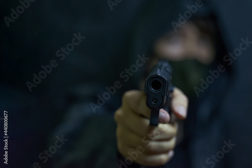 Man in hood and mask threatens with firearm. Revolver in person's hands. Murderer or armed thief. Criminal with a gun.