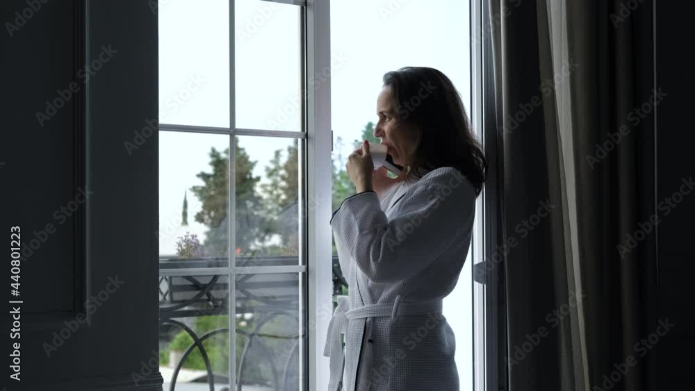 Beautiful Young Woman in a Bathrobe Talking by the Phone and Drinking Coffee while Standing near the Big Window of a Hotel Room or an Apartment. Travel, Tourism, Vacation and People Concept Beautiful Young Woman in a Bathrobe Talking by the Phone and Drinking Coffee while Standing near the Big Window of a Hotel Room or an Apartment. Travel, Tourism, Vacation and People Concept