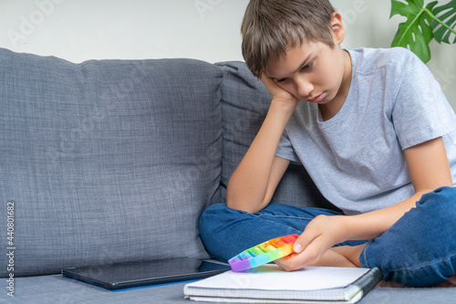 Tired unhappy teenager sitting with notebook and tablet computer and holding pop it fidget toy at home. Education, emotions, learning difficulties