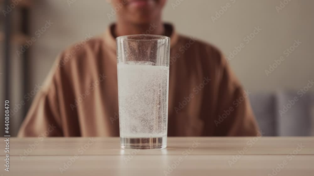 Close up sparkling water glass with dissolving effervescent aspirin pill standing on wooden table sick unhealthy dark skinned woman taking emergency medicine painkiller to relieve headache or fever. Close up sparkling water glass with dissolving effervescent aspirin pill standing on wooden table sick unhealthy dark skinned woman taking emergency medicine painkiller to relieve headache or fever.