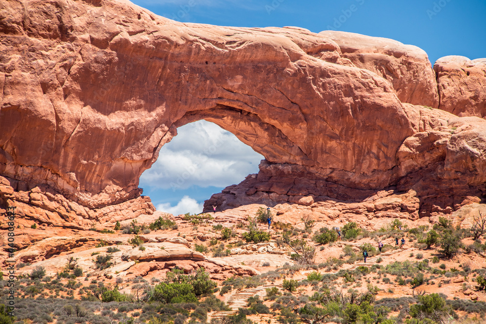 Huge natural red arch at Arches National Park in Utah USA with tourists walking up rock stairs toward it.