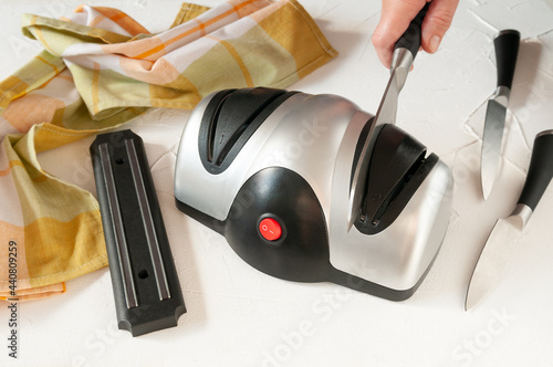 Electric knife sharpener. The plastic body is gray-black. In the sharpener there is a kitchen knife held by a woman's hand. Nearby are kitchen knives. Light background. photo