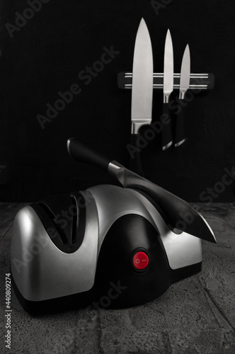 Electric knife sharpener. The plastic body is gray-black. A kitchen knife is inserted into the sharpener. In the background there are kitchen knives on the back. Dark concrete background. Close-up. photo