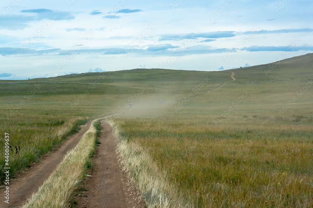 A steppe with grass and a road along which two cars drive in dust. Sky with clouds. Horizontal image.