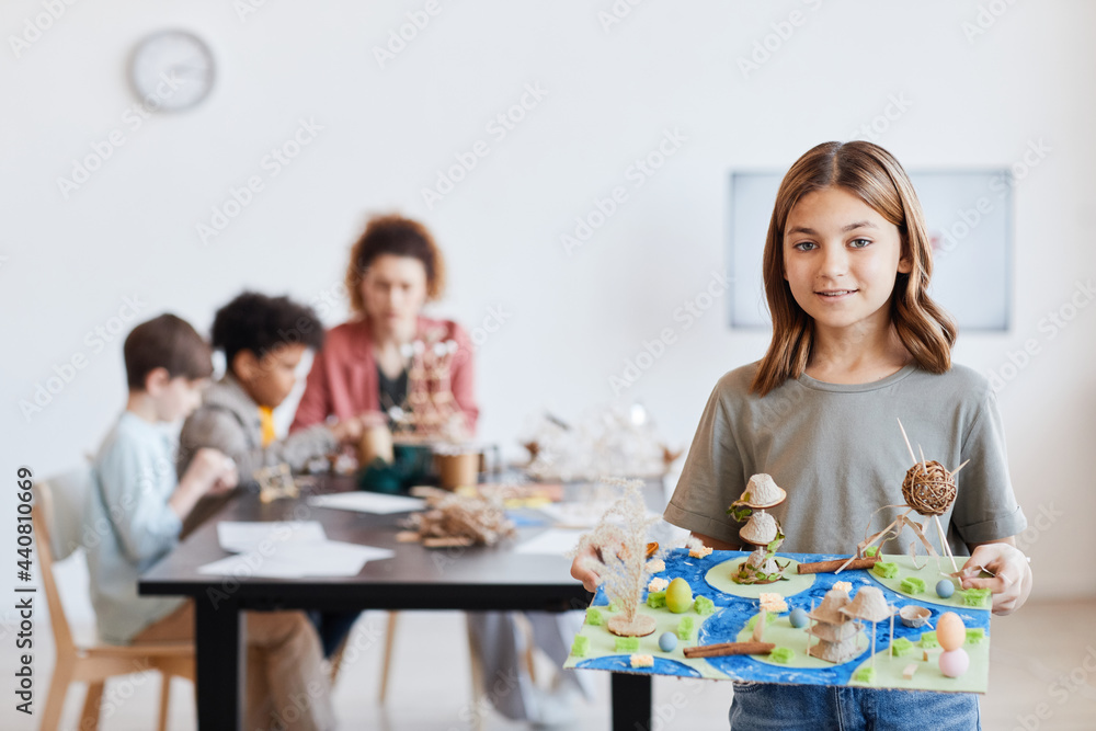 Waist up portrait of smiling schoolgirl holding model and looking at camera during art and craft class in school, copy space Waist up portrait of smiling schoolgirl holding model and looking at camera during art and craft class in school, copy space