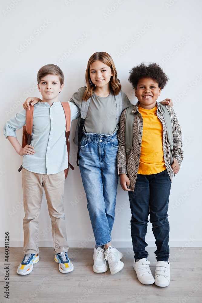 Full length portrait of three smiling schoolkids wearing backpacks and looking at camera while standing against white wall Full length portrait of three smiling schoolkids wearing backpacks and looking at camera while standing against white wall