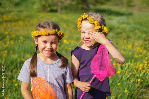 Two little girls with wreaths of yellow dandelions and nets in their hands stand against the background of a green meadow covered with dandelions  frown from the sun  one covers her face with her hand