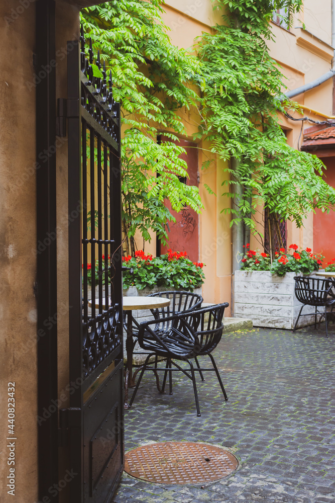 Cozy street cafe with iron fence in old town. Empty patio with outdoor furniture and red flowers. Hotel terrace with chairs and tables. Sidewalk cafe decoration in backyard. Beautiful outdoor cafe.