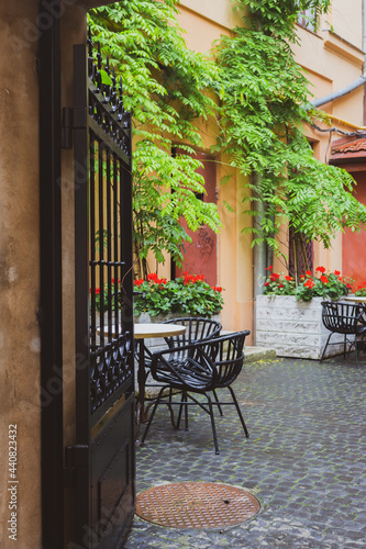 Cozy street cafe with iron fence in old town. Empty patio with outdoor furniture and red flowers. Hotel terrace with chairs and tables. Sidewalk cafe decoration in backyard. Beautiful outdoor cafe.