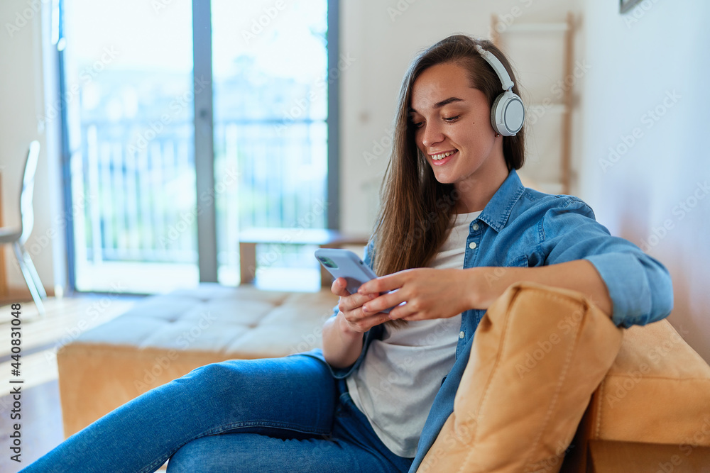 Smiling casual happy young girl sitting on a sofa and using smartphone and wireless headphone for watching video content online Smiling casual happy young girl sitting on a sofa and using smartphone and wireless headphone for watching video content online