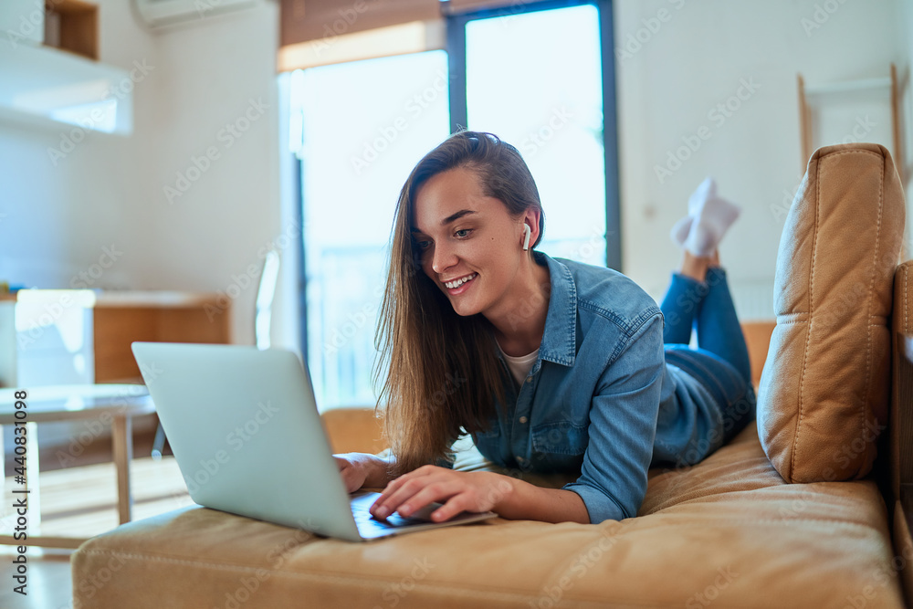 Fototapeta premium Casual smart modern smiling millennial girl with wireless headphone using laptop for watching video content, checking social media and shopping online during relax on sofa at home .