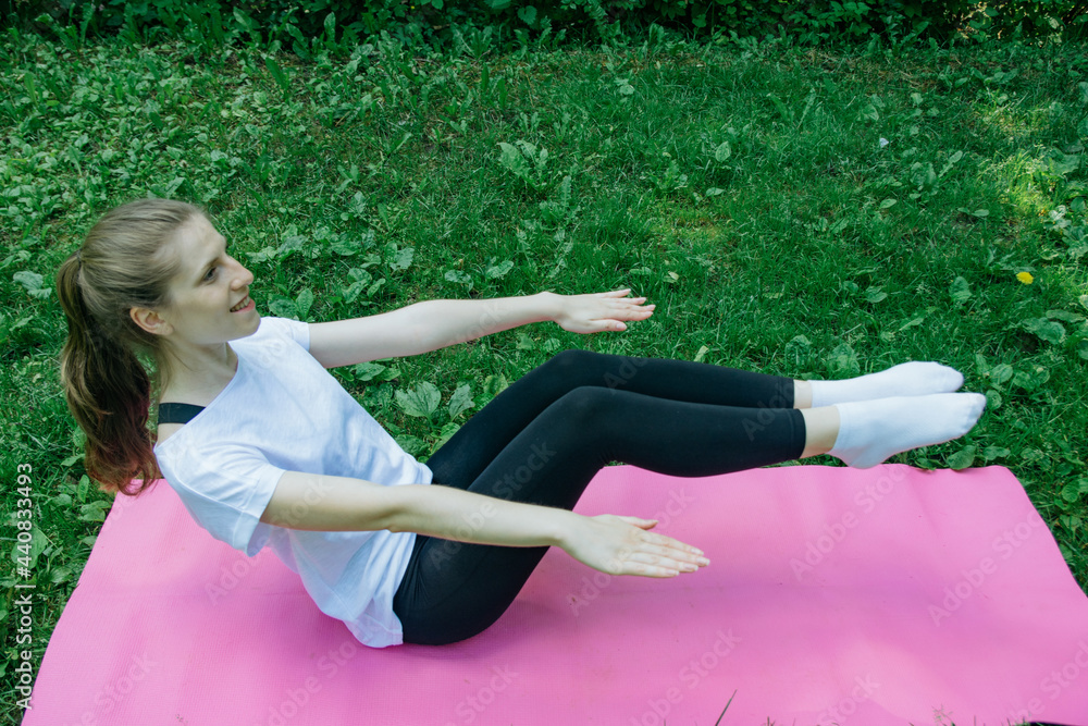 Fototapeta premium A girl in a white T-shirt and black leggings on a pink mat on a summer green lawn and shakes the press