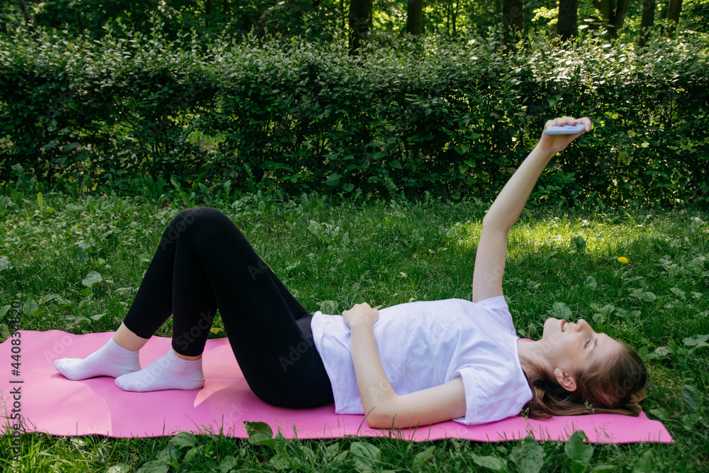 Fototapeta premium A girl in a white T-shirt and black leggings on a pink mat on a summer green lawn and talking by video call on the phone