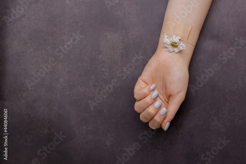 Close-up of woman's hand with flowers attached to her forearm with adhesive bandage on a dark fabric background. Healing or recovering concept. Gray manicure. concept of mental health, medical care.