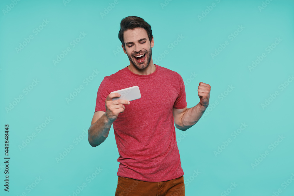 Handsome young smiling man in casual clothing using smart phone while standing against blue background Handsome young smiling man in casual clothing using smart phone while standing against blue background
