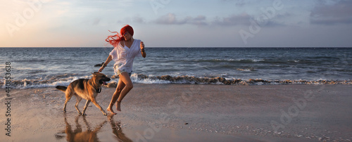 Woman with long dyed red hair running with her dog on a beach. Friendship, fun and enjoyment at summer vacation