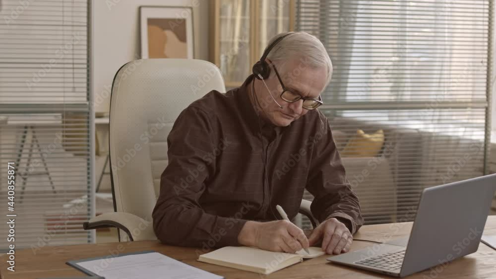 Medium slowmo of successful senior businessman in smart casual clothes wearing headset while talking to business partners and taking notes sitting by work desk in modern office Medium slowmo of successful senior businessman in smart casual clothes wearing headset while talking to business partners and taking notes sitting by work desk in modern office