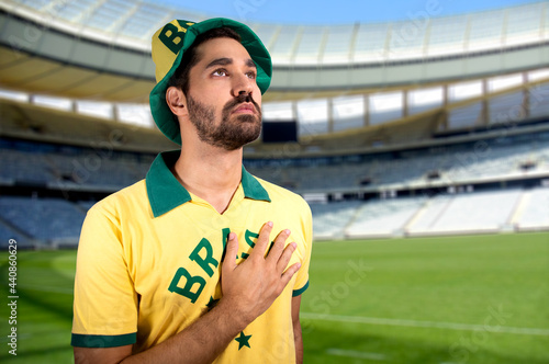 Brazilian sports fan in a position of respect to hear the national anthem, wearing a green and yellow hat. Football stadium in the background.