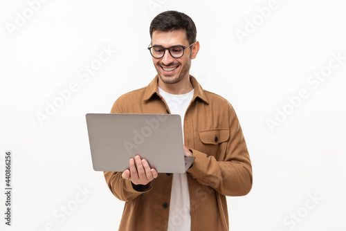 Young happy man standing with opened laptop, browsing online or typing message, isolated on gray background