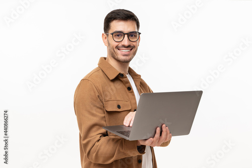 Portrait of young modern business man standing in casual brown shirt, holding laptop and looking at camera with happy smile, isolated on gray photo