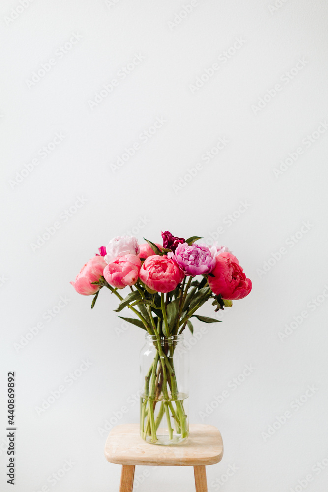 A bright image of a bouquet of flowers in a clear vase against a white wall. The peony flowers are cheerful in colors of pink, red, magenta, and violet.