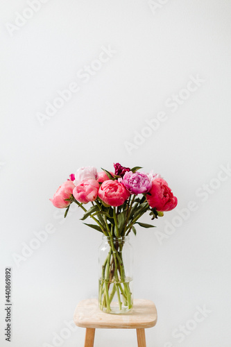 A bright image of a bouquet of flowers in a clear vase against a white wall. The peony flowers are cheerful in colors of pink  red  magenta  and violet.