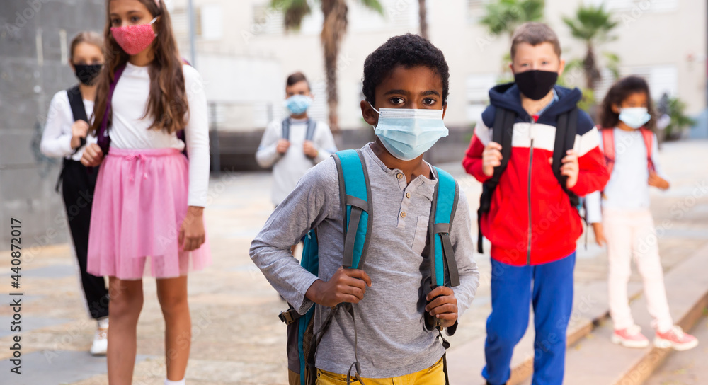 Portrait of schoolboy in medical mask standing near school, kids on background