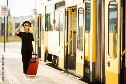 Young casual female traveler in total black and sunglasses walking and pulling a red wheeled luggage. Concept of travel. Copy space