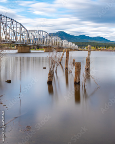 Nisutlin Bay Bridge in Teslin  Yukon Territory  northern Canada with calm water  old wooden posts in the river from the shore. Mountains in the background on summer spring day with steel structure. 