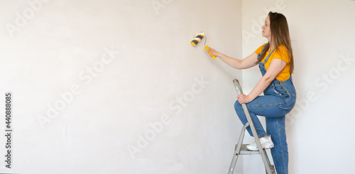 A pensive woman with roller and ladder looks dreamily in a room with an unpainted wall with copy space. Independent single female makes DIY repairs in her apartment